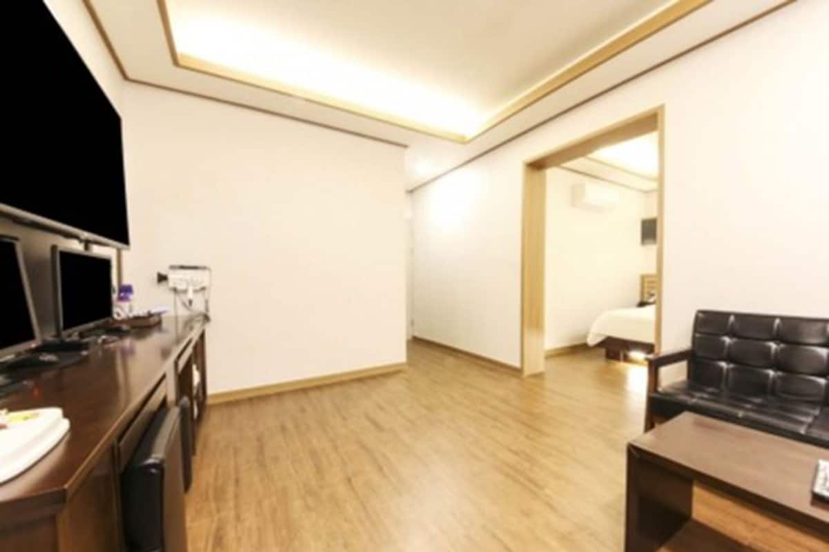 Habitación de hotel moderna con piso de madera, sofá de cuero negro, una mesita de centro y un televisor sobre un mueble de madera.