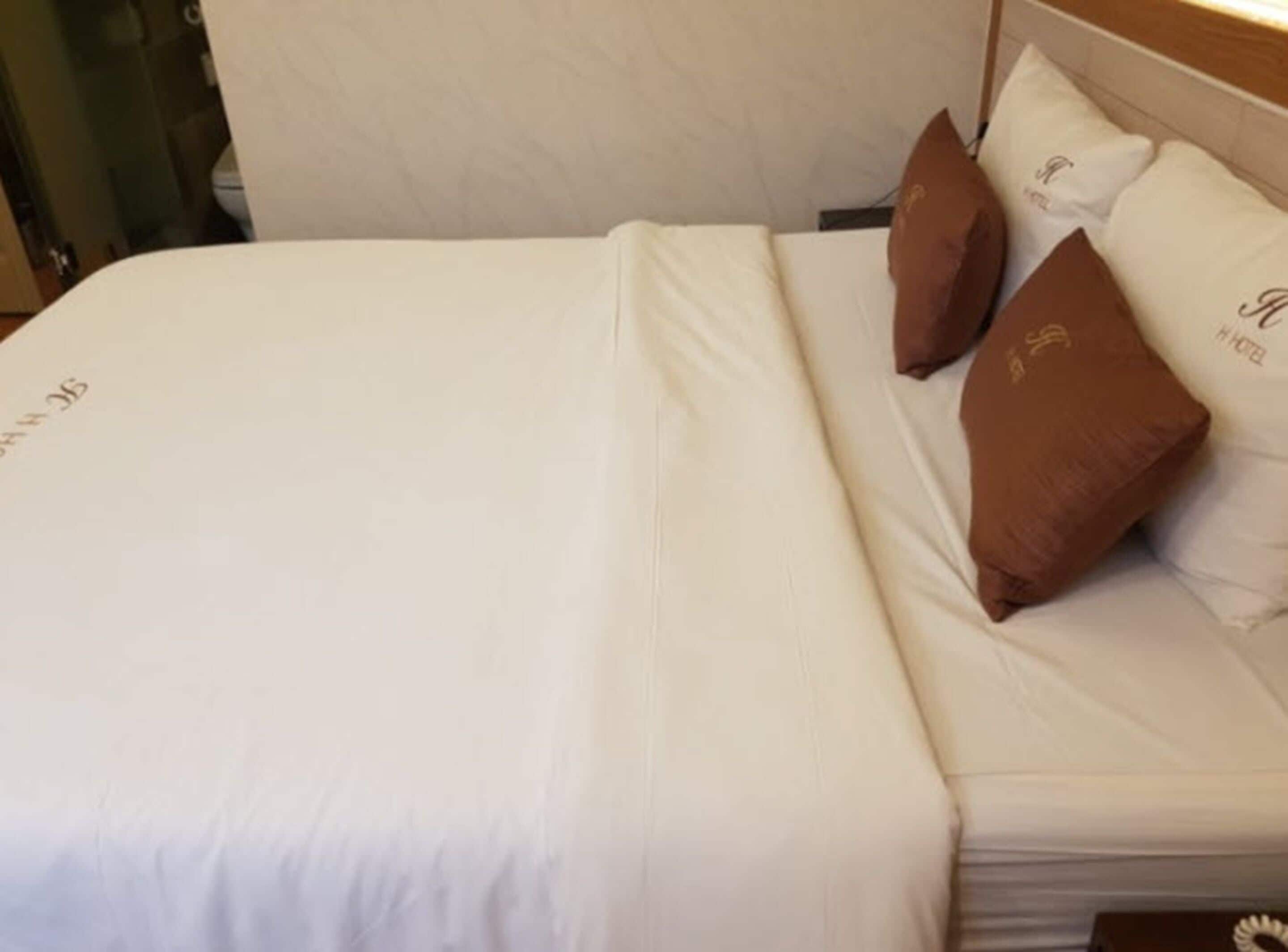 A neatly made bed with white bedding and three brown pillows, one of which has a logo.