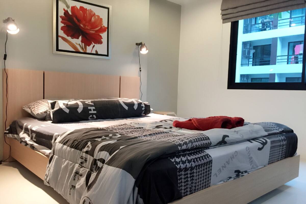 A neatly made bed with patterned bedding, a red throw blanket, and a framed floral artwork above it.