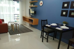 A modern living room with a dining area, a TV mounted on a wooden shelf, and a balcony visible through the window.