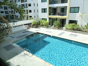 A swimming pool with tiled flooring, surrounded by potted plants and a building with balconies.