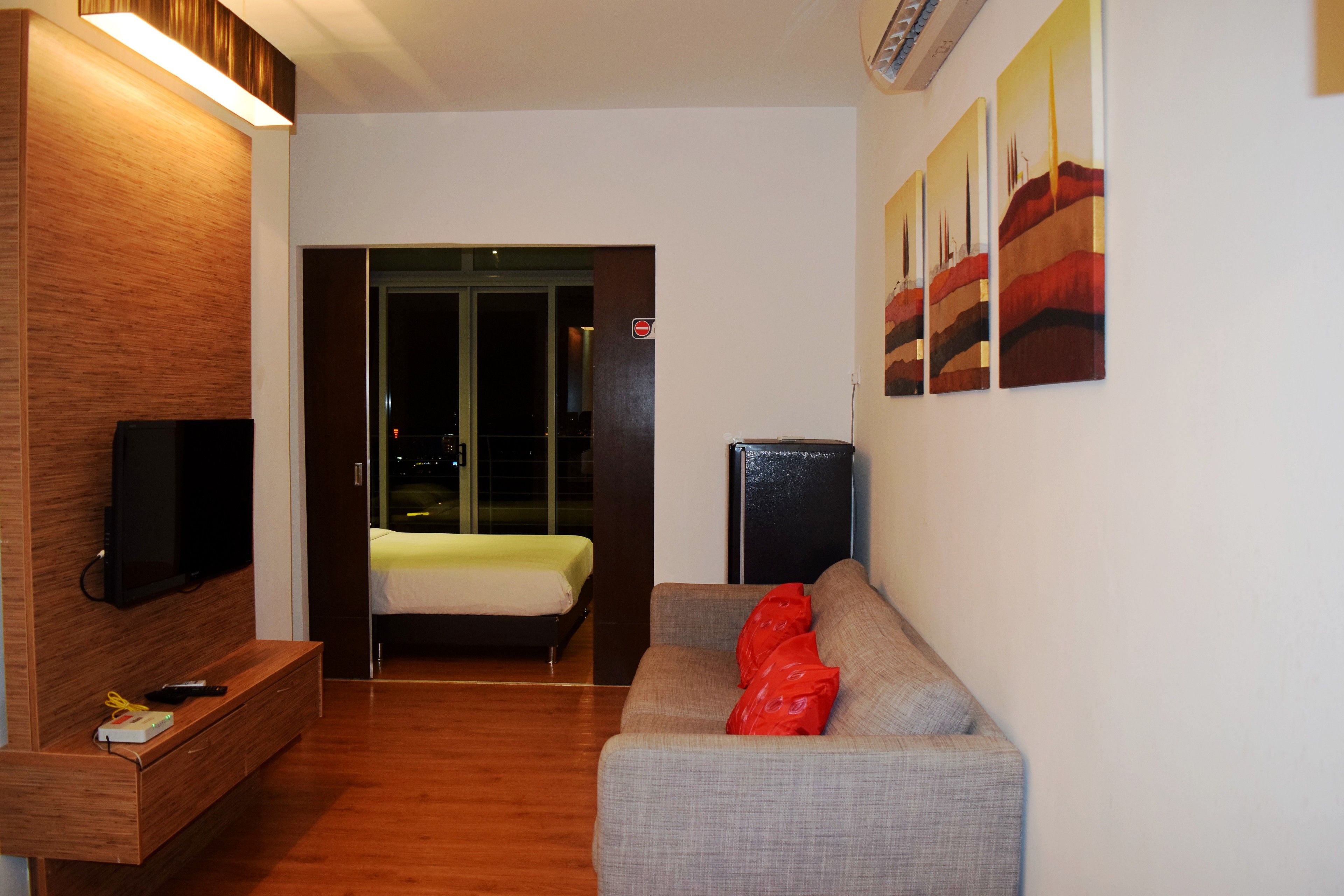A modern hotel room with a wooden accent wall, a flat-screen TV, a grey sofa with red cushions, a bed with a yellow bedspread, and a small black speaker.