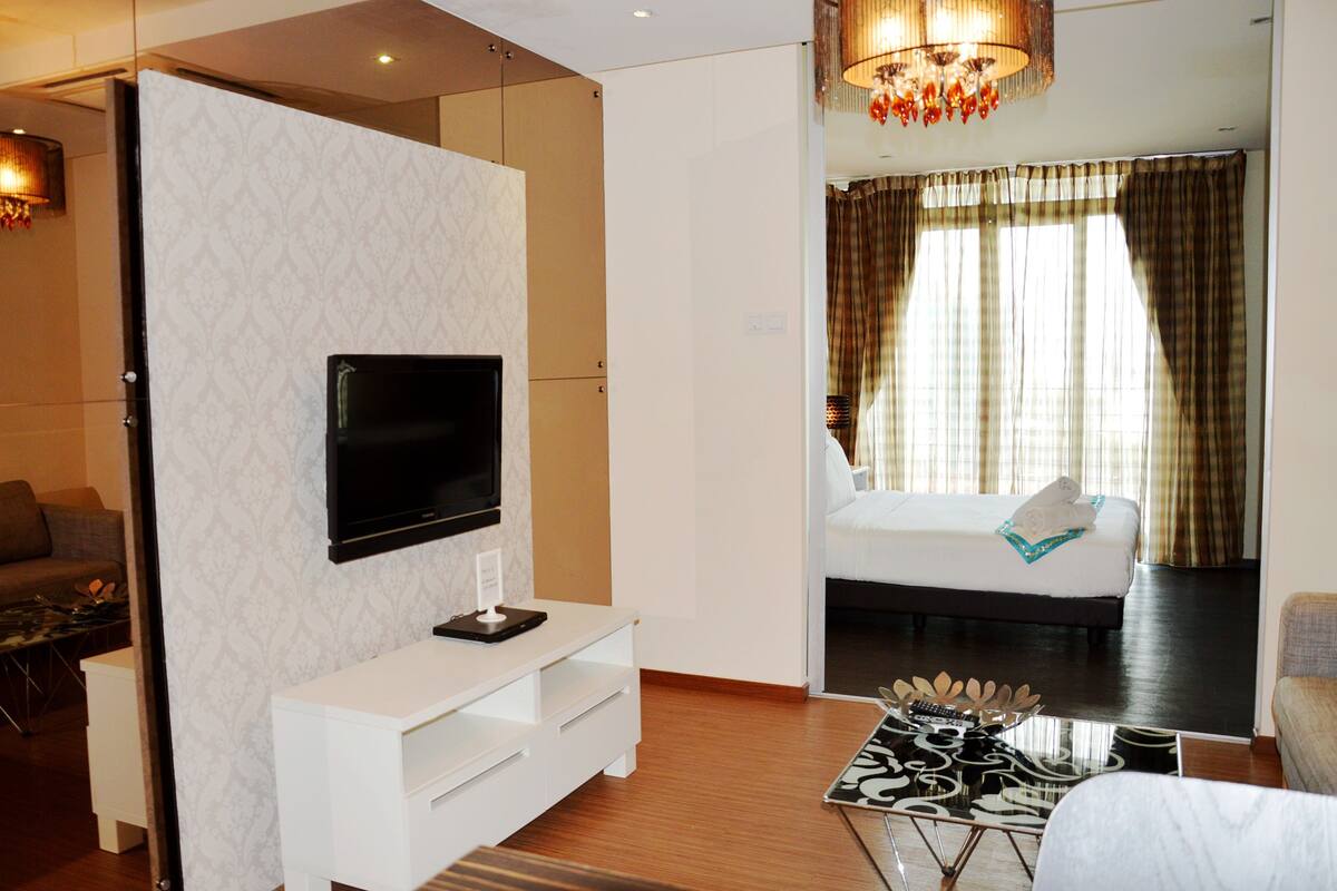 Una habitación de hotel moderna con una televisión de pantalla plana montada en una pared blanca, una cama con cabecera blanca, una mesa de comedor con camino de mesa blanco y negro, y un ventanal con cortinas.