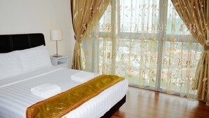 Una cama bien hecha con sábanas blancas y un detalle dorado, una mesita de noche con lámpara y cortinas traslúcidas con estampado floral.