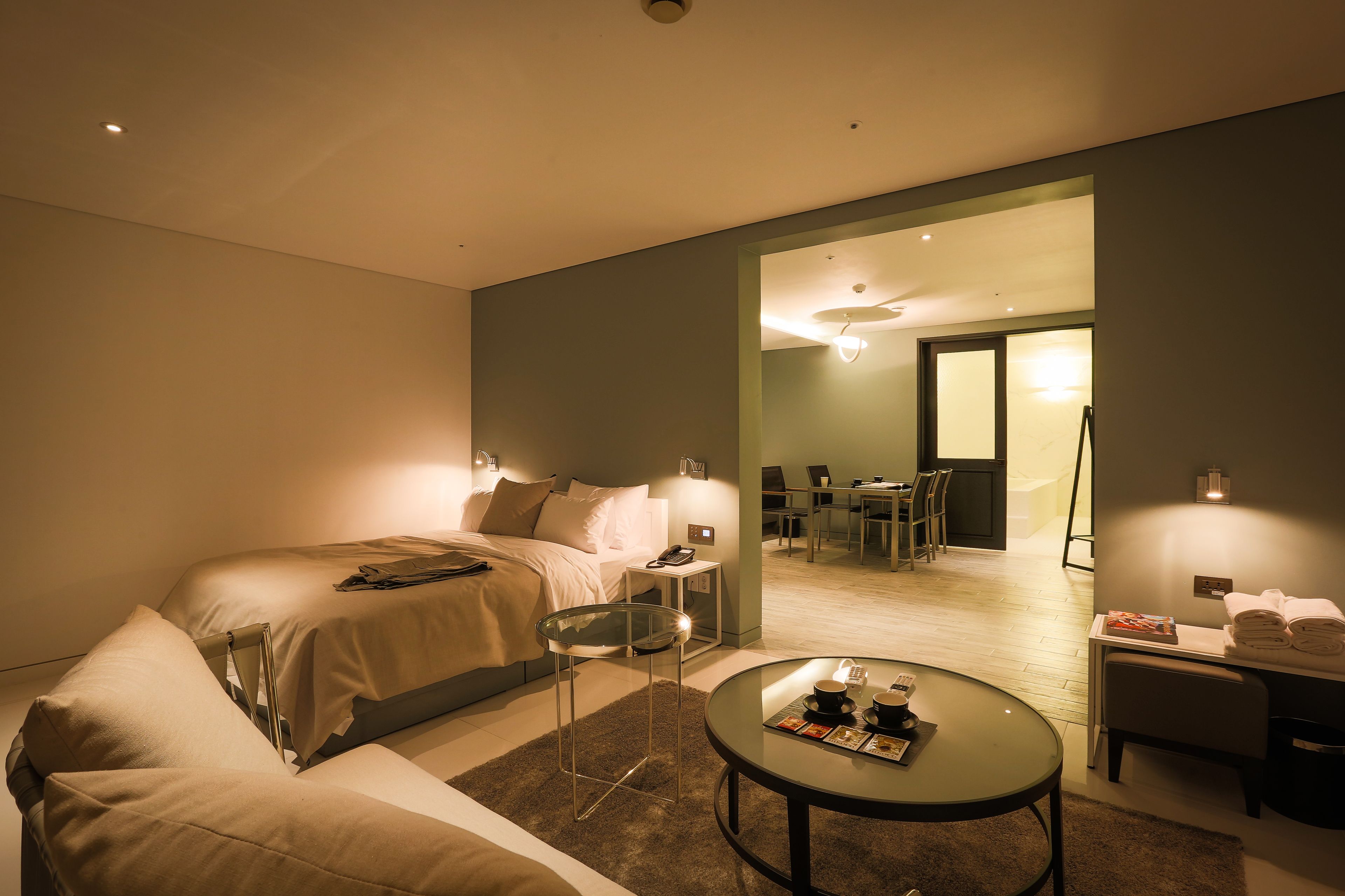 A hotel room with a bed, a glass table with magazines, a chair, a desk with a lamp, and a bathroom visible through an open door.