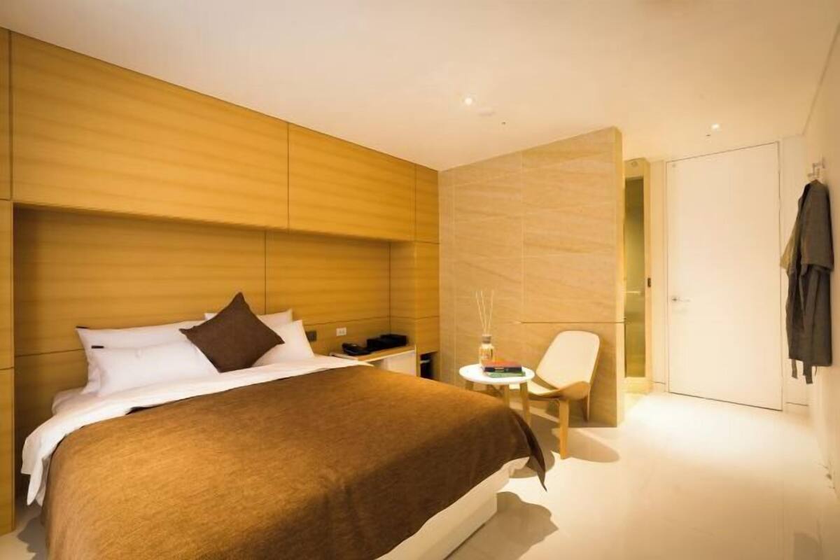A modern hotel room with a large bed, a small table with a chair, and a wardrobe.