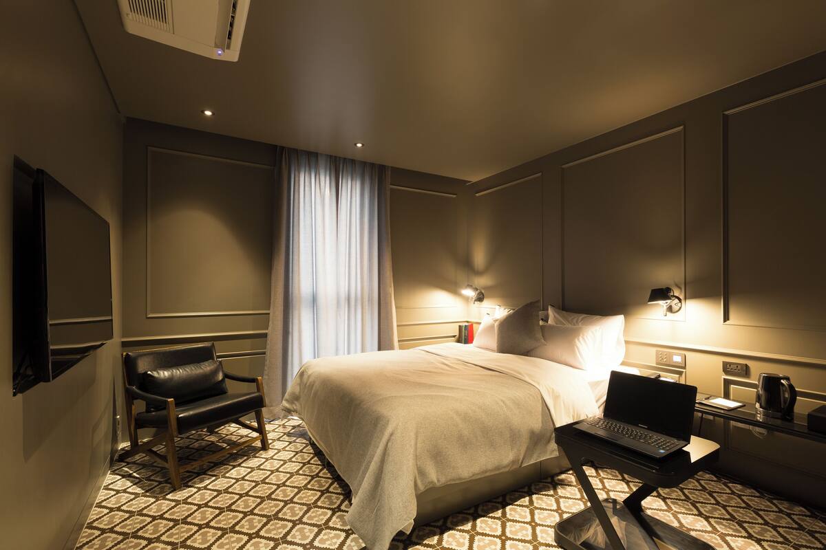 A hotel room with a bed, a desk with a laptop, a chair, a bedside table with a lamp, and a large window with curtains.