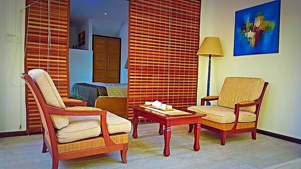 A room with two wooden chairs, a small table, and a lamp. There is a painting on the wall and a window with blinds.
