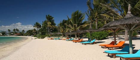 A beach with white sand, palm trees, and rows of orange and blue lounge chairs under thatched umbrellas.