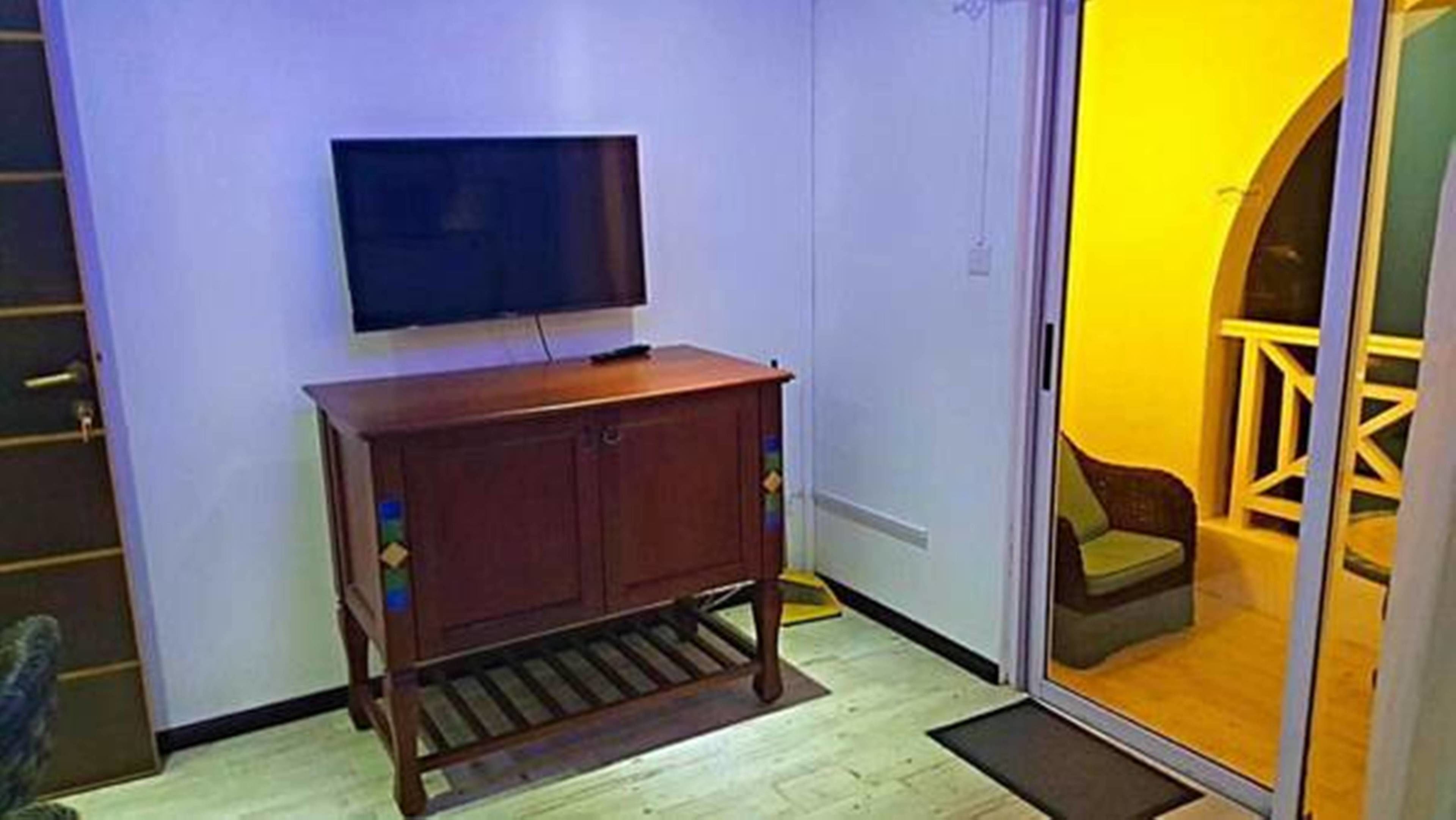A room with a wooden cabinet, a flat-screen TV, and a sliding glass door leading to an outdoor area with a yellow-lit seating area.