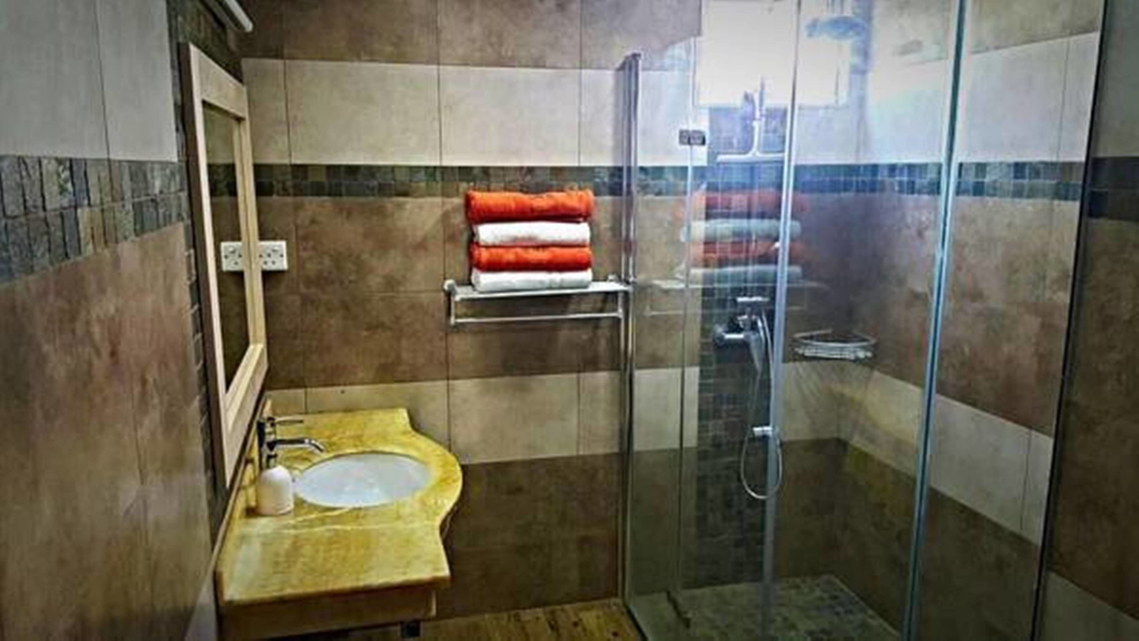 Een badkamer met een douche met glazen wand, een wastafel met een geel aanrecht en een handdoekrek met opgevouwen handdoeken.
