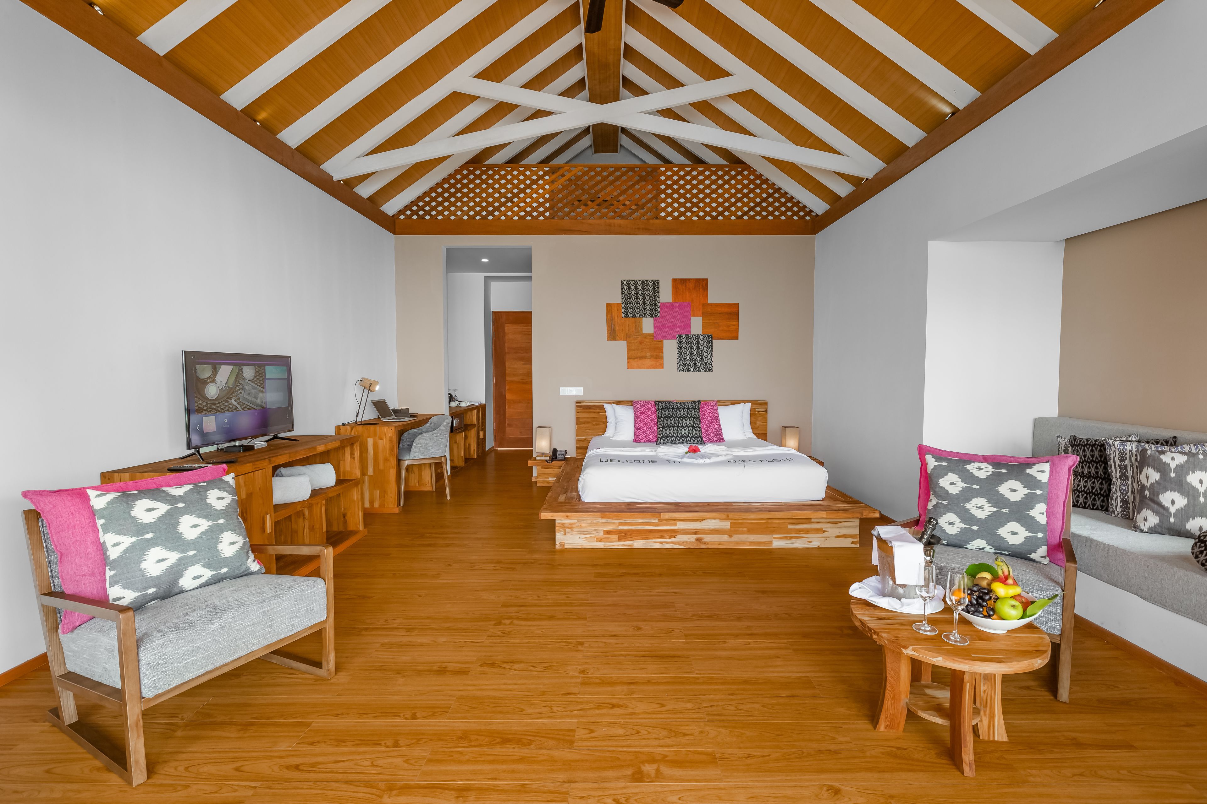 Una habitación amplia con techo de madera, una cama con colcha blanca y rosa, un escritorio de madera con una computadora y un sofá con cojines estampados.
