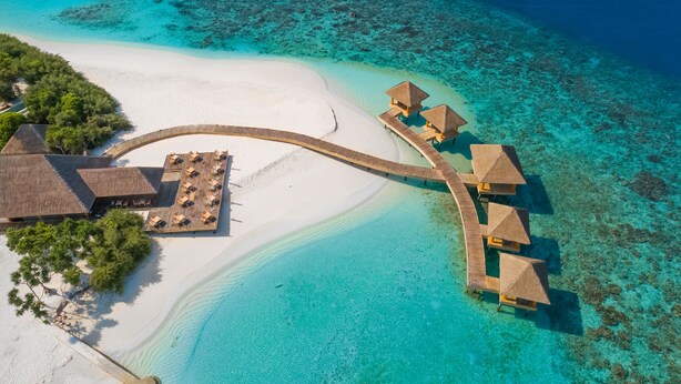 Vue aérienne d’un complexe hôtelier avec des bungalows sur pilotis reliés par une passerelle en bois, une plage de sable et des eaux turquoise cristallines.
