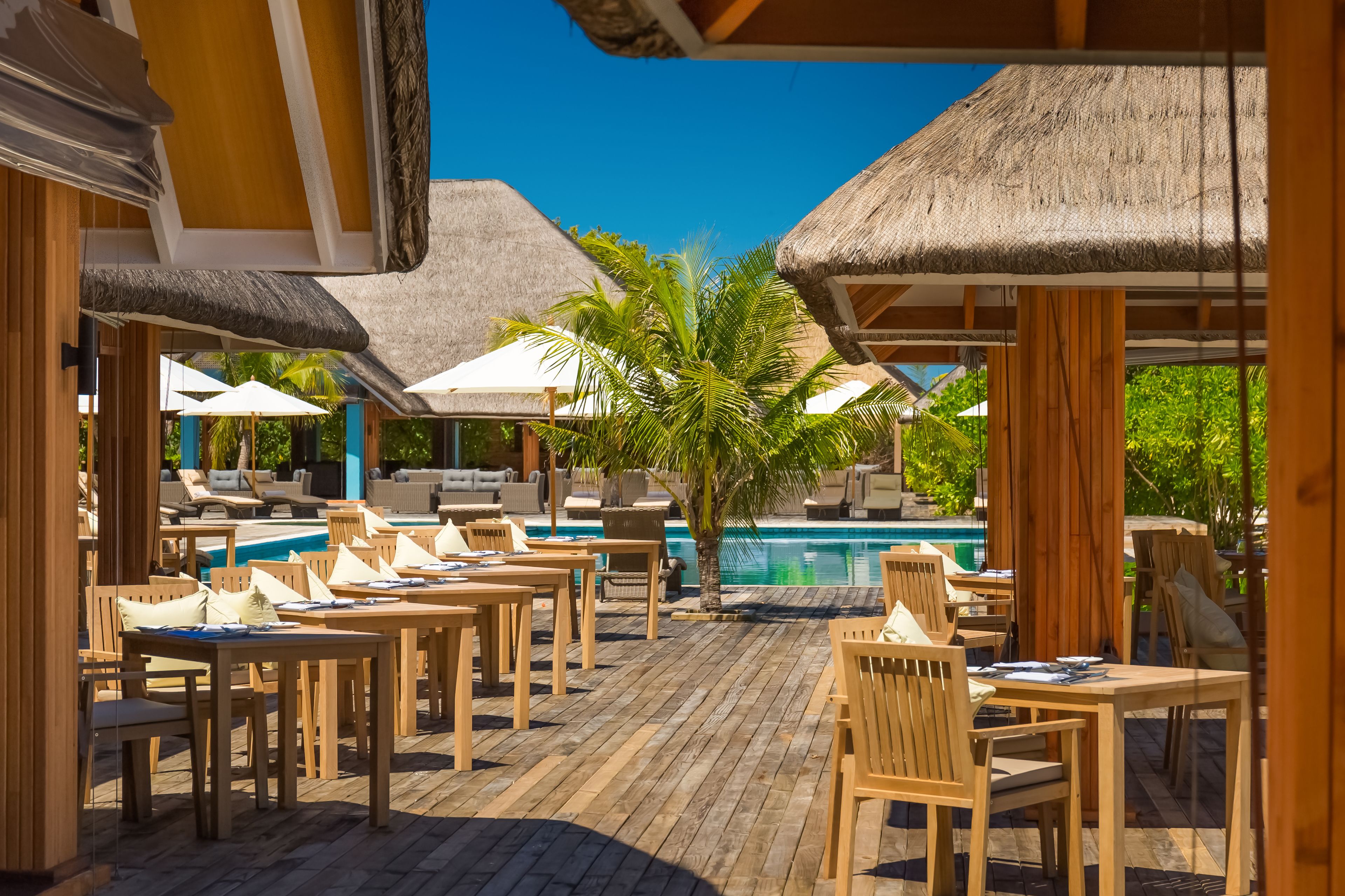 A poolside area with wooden tables and chairs, thatched umbrellas, and a clear blue sky.