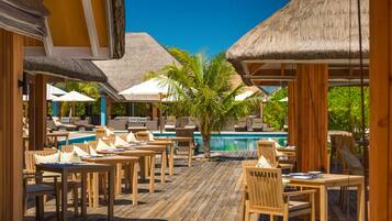 A poolside area with wooden tables and chairs, thatched umbrellas, and a clear blue sky.