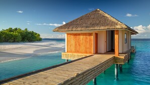 Un bungalow en bois sur pilotis, doté d’un toit de chaume, relié à une terrasse en bois qui s’étend sur une eau turquoise et limpide, et entouré d’une végétation luxuriante et d’une plage de sable.