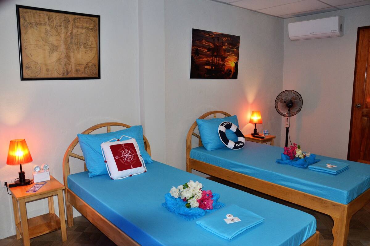 A room with two beds, each with a blue bedspread and pillows, a bedside table with a lamp, and a wall-mounted air conditioner.