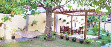 A covered patio area with a hammock, wooden chairs, and potted plants.