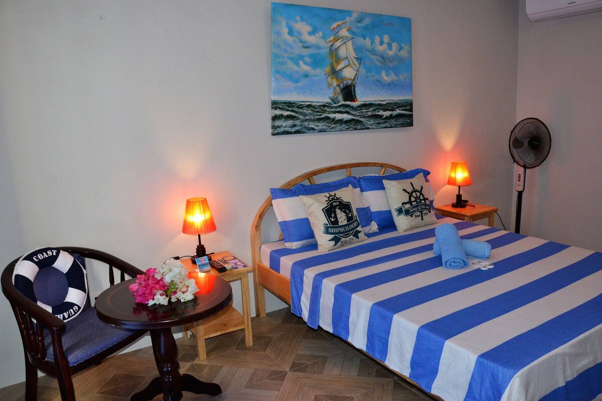 A room with a bed, a chair, a table, and a painting of a ship on the wall.