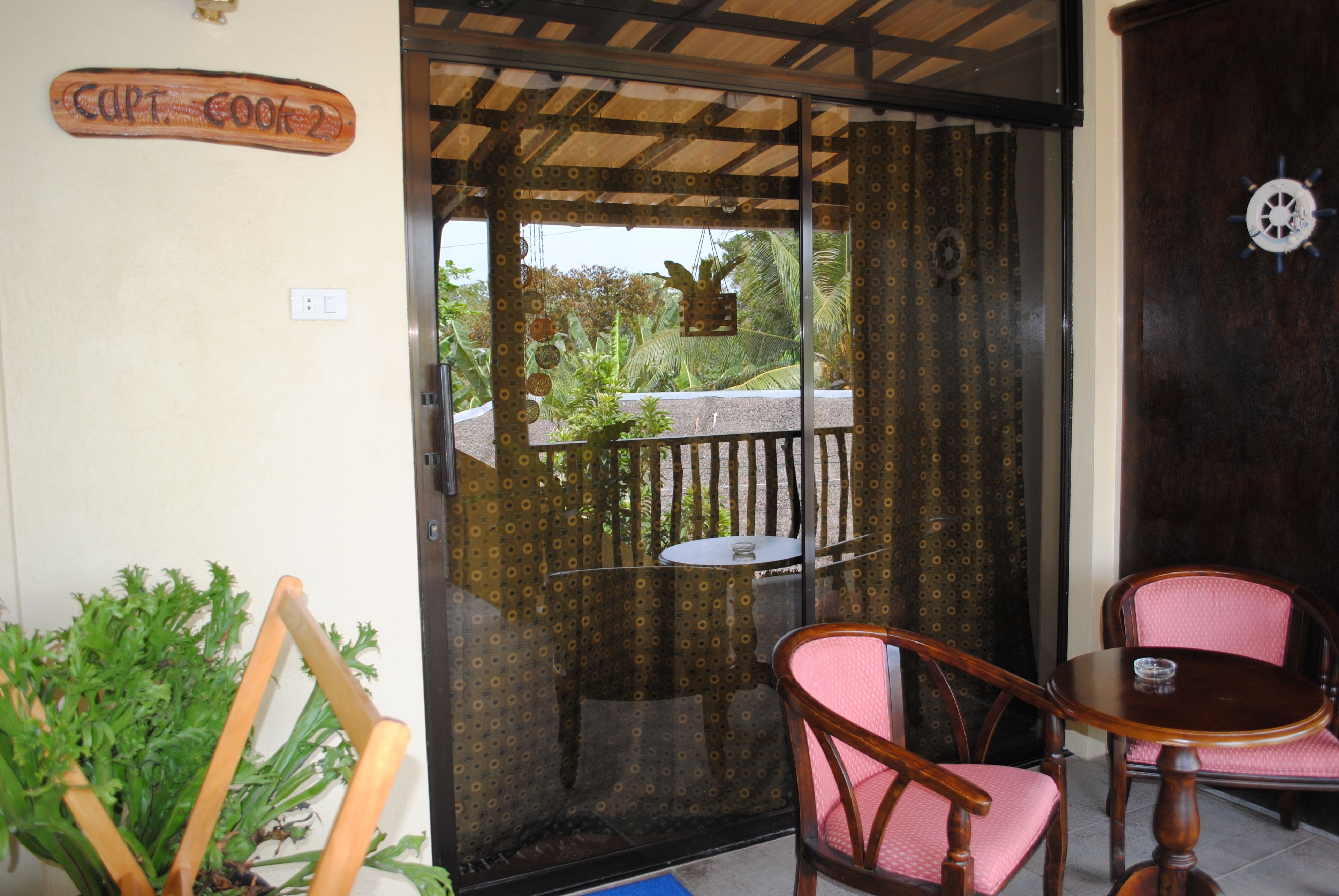 A room with a glass door leading to a balcony, a small table, two chairs, and a sign reading "Capt. Cook 2".