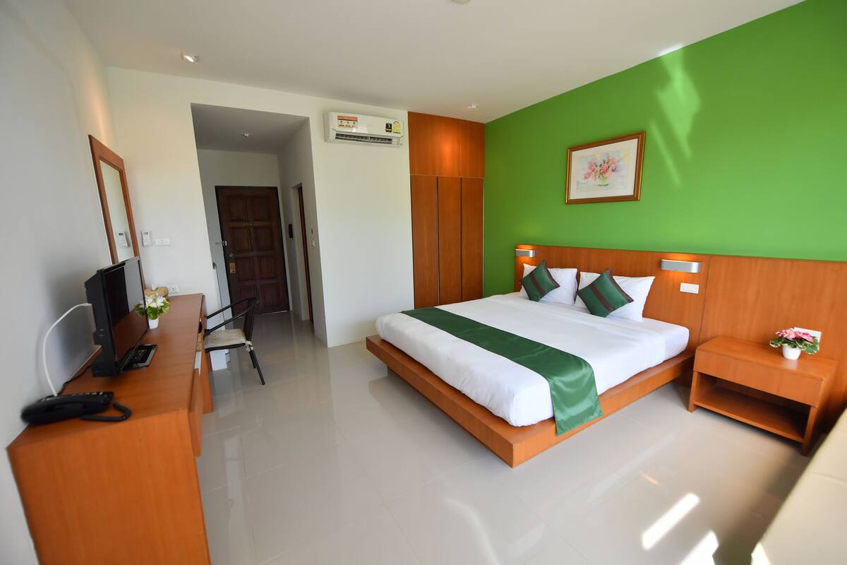 A hotel room with a bed, a desk with a computer, a telephone, and a small bedside table with a flower.