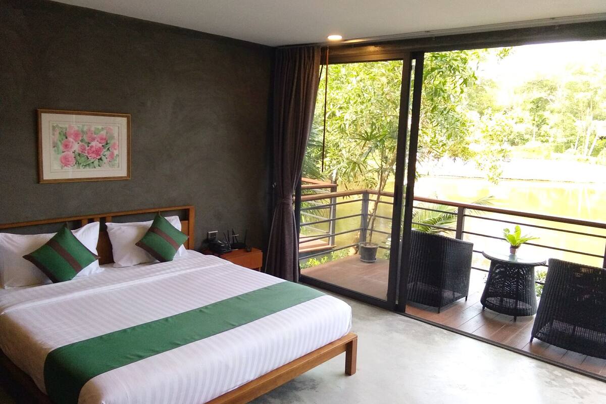 A hotel room with a large bed, a small table, a chair, and a balcony with a view of greenery and a body of water.