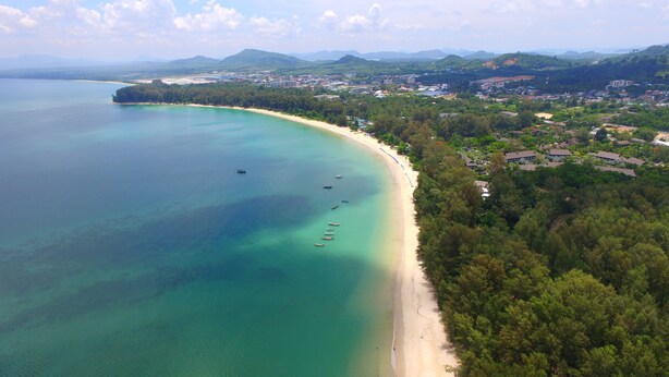 An aerial view of a coastal area with a sandy beach, clear turquoise waters, and a dense forested area with buildings. - The Touch Green Naiyang Hotel & Fitness (Sa Khu)