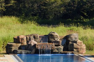 Una cascada de roca en una piscina con diseño natural, rodeada de hierba y árboles.