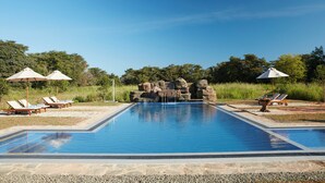 A rectangular swimming pool with a waterfall feature, surrounded by lounge chairs and umbrellas, set against a backdrop of trees and a clear sky. - Seerock The King's Domain (Avudangawa)