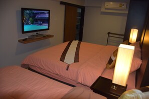 Habitación de hotel con televisión en un estante, cama con ropa de cama rosa, mesita de noche con lámpara y aire acondicionado instalado en la pared.