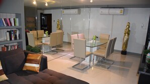 A modern living room with a sofa, a glass dining table, and chairs. There is a bookshelf with books and decorative items. - Kalim Residence Unit - 215 (Patong)