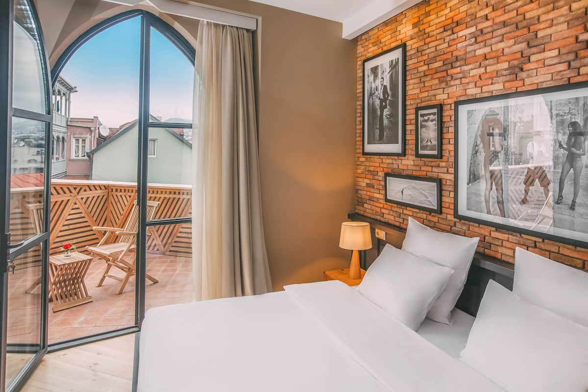 A hotel room with a large bed, a wooden chair, a small table with a flower, a lamp, and a balcony with a view of buildings.