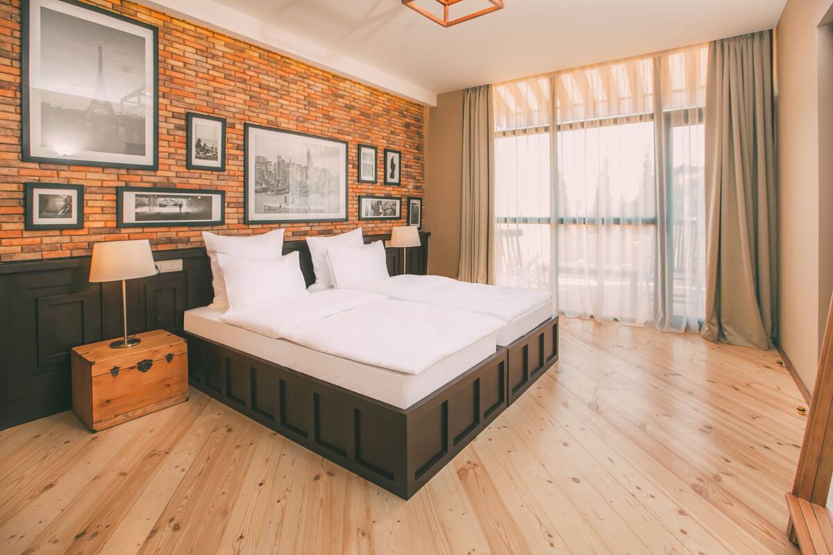 A modern hotel room with a brick-patterned wall, a large bed, a wooden nightstand, and a window with curtains.