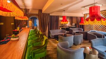Un área de bar moderna con taburetes verdes, una barra de madera y lámparas colgantes rojas. Hay sillones azules y una pequeña mesa con un jarrón de flores.