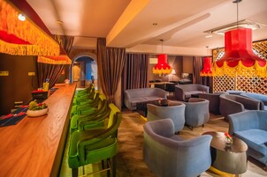 Un área de bar moderna con taburetes verdes, una barra de madera y lámparas colgantes rojas. Hay sillones azules y una pequeña mesa con un jarrón de flores.