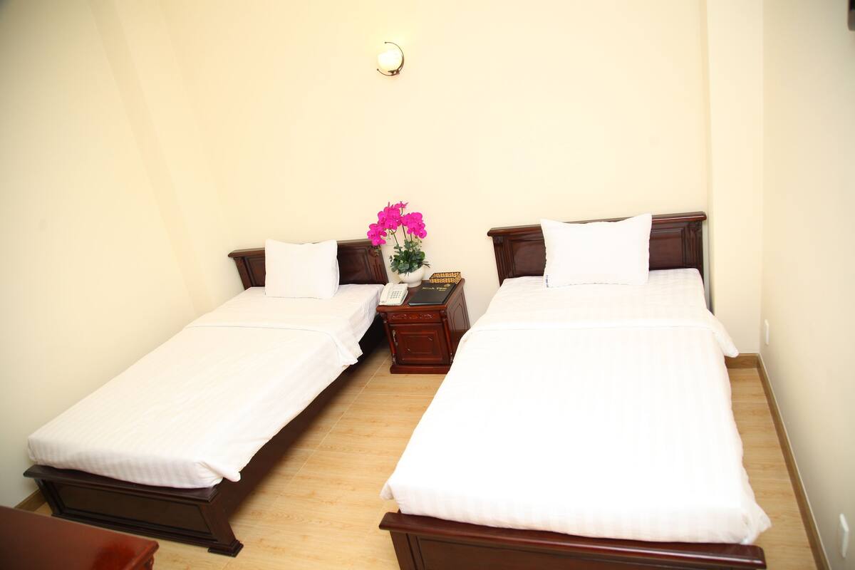 A hotel room with two single beds, a nightstand with a telephone, and a wall-mounted light fixture.