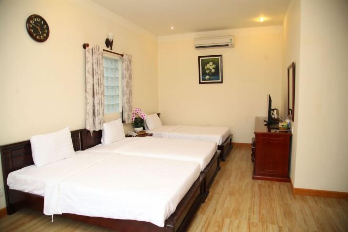 A hotel room with two single beds, a wooden floor, a clock, an air conditioner, and a small desk with a mirror.