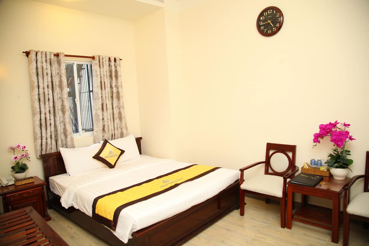 A neatly arranged hotel room with a bed, a bedside table with a lamp, a chair, and a small table with a vase of flowers.