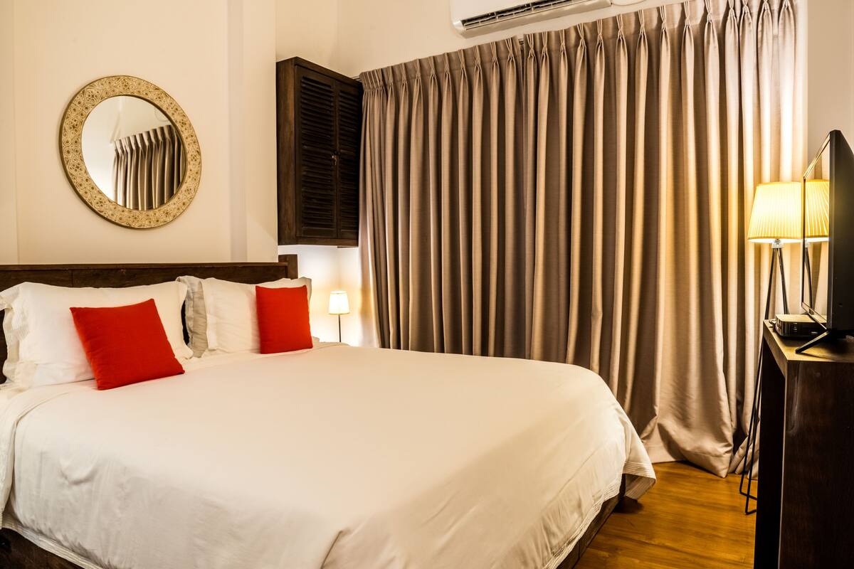A hotel room with a large bed, a round mirror on the wall, a television on a stand, and a window with curtains.