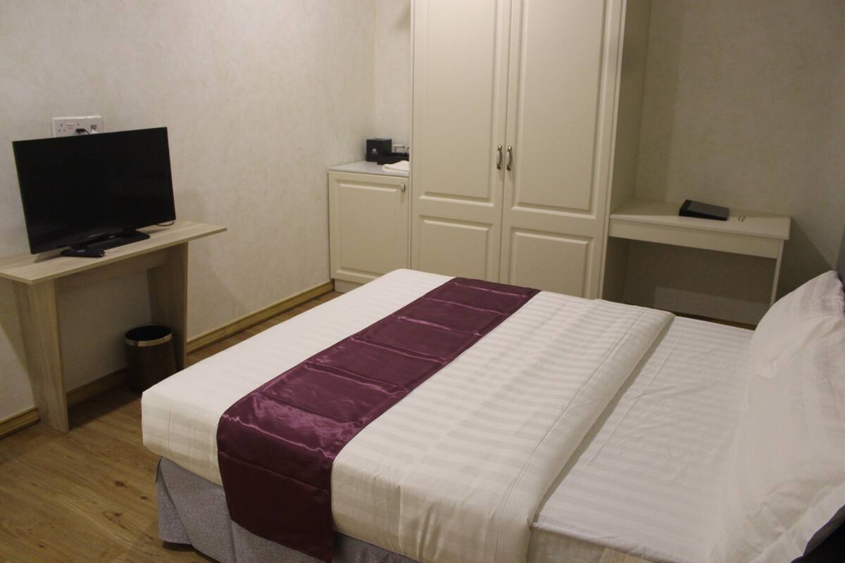 A hotel room with a bed, a television on a desk, a wardrobe, and a small shelf.