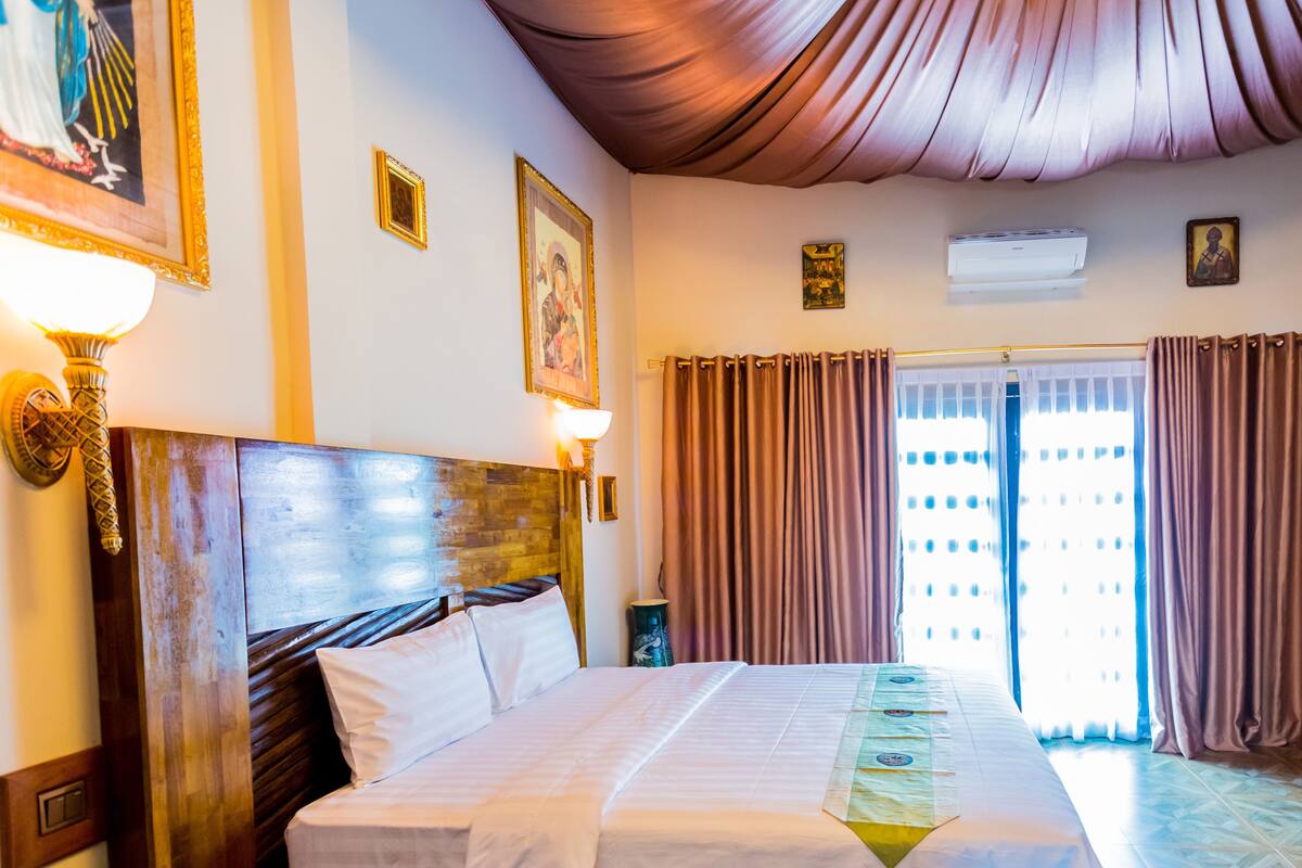 A hotel room with a large bed, a wooden headboard, a wall-mounted lamp, and a window with curtains.