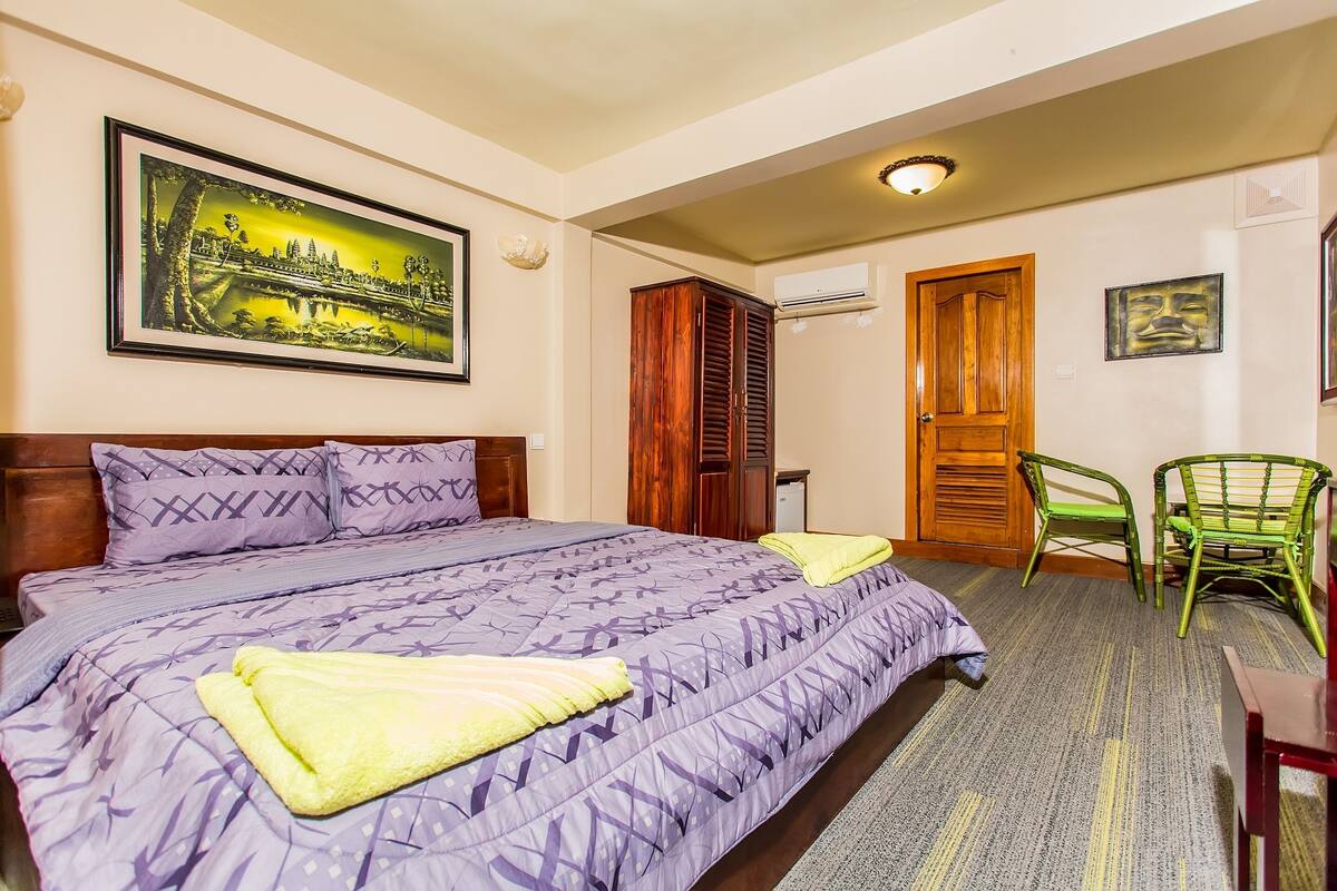 A hotel room with a large bed, a wooden wardrobe, a green chair, and a painting on the wall.