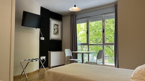 A hotel room with a bed, a desk, a chair, a television, and a window with a view of trees. - Apartamentos Leon (León)