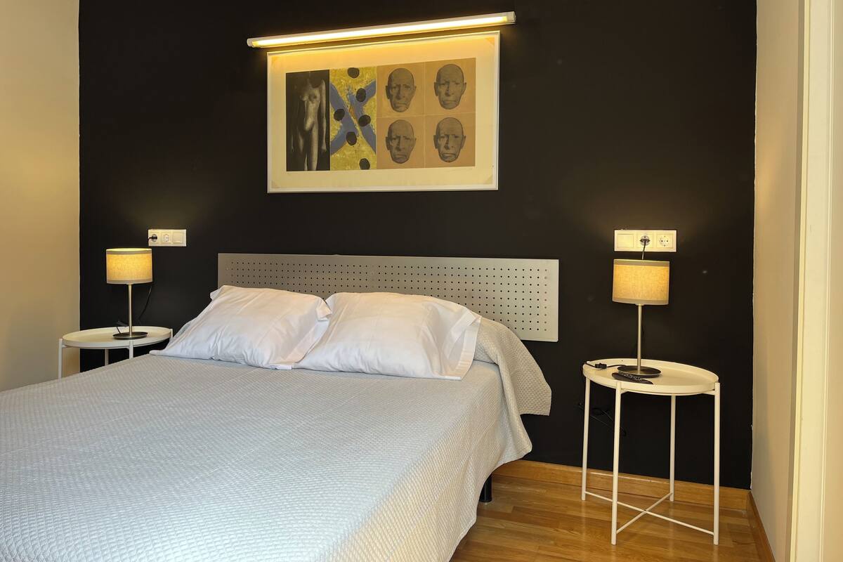 A neatly made bed with white linens, two bedside tables with lamps, a headboard with a perforated design, and a framed artwork above the bed.