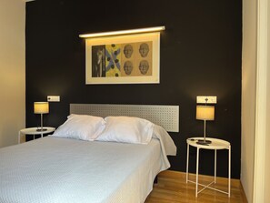 A neatly made bed with white linens, two bedside tables with lamps, a headboard with a perforated design, and a framed artwork above the bed. - Apartamentos Leon (León)