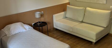Habitación de hotel con una cama, un sofá blanco, una mesita de noche con una lámpara y un cuadro en la pared.