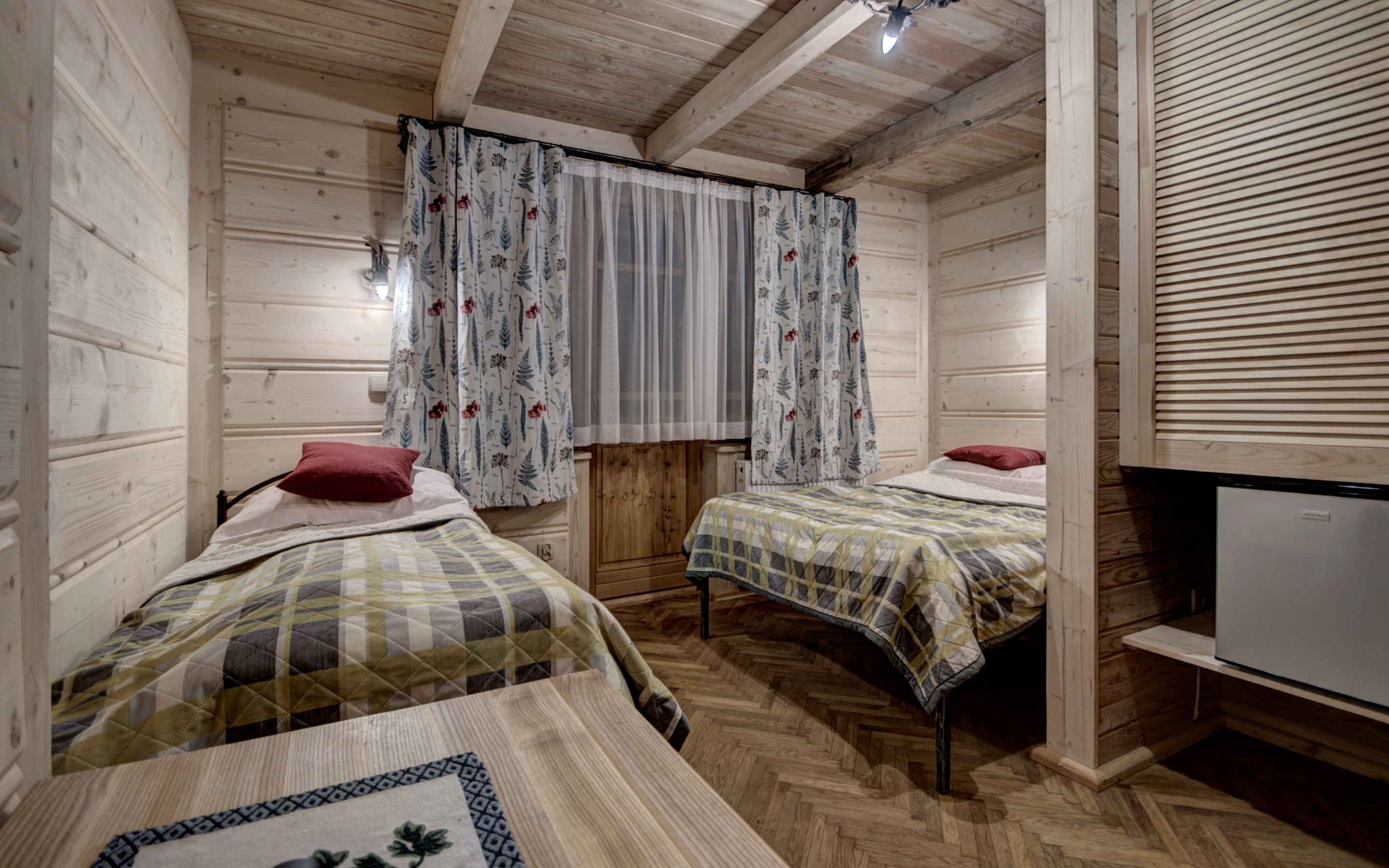 A cabin-style room with two beds, wooden walls, a window with floral curtains, and a small refrigerator.