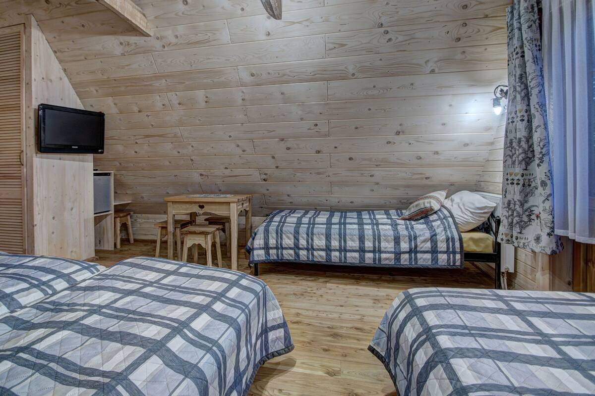 Una habitación de cabaña acogedora, con paredes y suelos de madera, dos camas con ropa de cama escocesa, una mesita pequeña, un televisor montado en la pared y una ventana con cortinas.