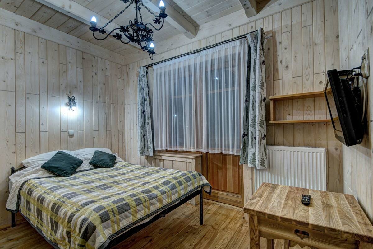 Un dormitorio con paneles de madera, una cama, una mesita, un radiador y un televisor montado en la pared.