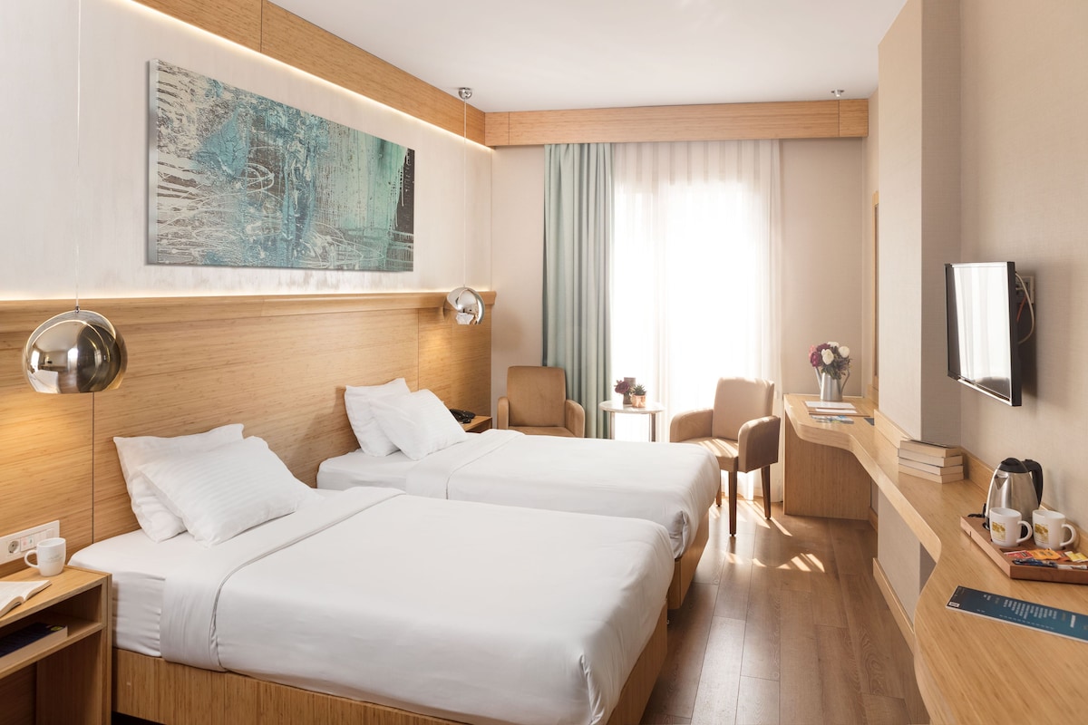 A hotel room with two beds, a desk, a chair, a TV, and a painting on the wall.